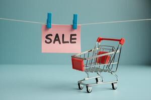 Miniature shopping cart with red handle, pink SALE note clipped on line, soft blue background photo