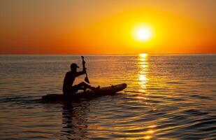Silhouette of a man floating on a board with an oar. Dawn sun on the sea. Swimming on a supboard. Water active sports. Beautiful landscape. Sunset on the sea. Ocean coast. Copy space. photo
