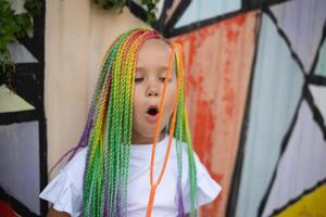 girl with multi-colored afro pigtails inflates soap bubbles against the background of a wall photo