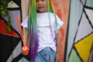 girl with multi-colored afro pigtails inflates soap bubbles against the background of a wall photo