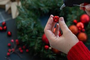 Crafter using hot glue Step by step instruction of making handmade Christmas wreath made of festive decorations in red color. Female hands creating craft homemade decor. New year celebration. Winter holidays Step 2 photo