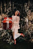 a girl in a Santa hat with a big Christmas gift in her hands on a fairy-tale island.Smiling woman in white clothes on the background of Christmas trees and small houses photo