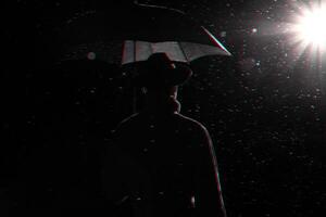 dark silhouette of a man in a raincoat and hat under an umbrella on the street in the rain photo
