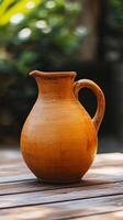 A rustic brown clay pitcher sits on a wooden table with a blurry green background. photo