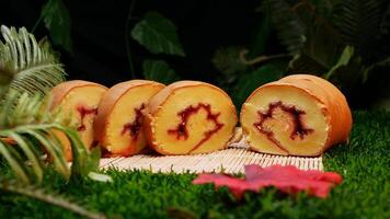 Four slices of rolled cake with red jam inside, placed on a bamboo mat and decorated with green leaves and fern plants. photo