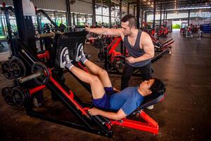 A male trainer is helping his trainee use a leg press machine by pushing the handles of the machine. photo