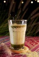 Matcha latte in a large glass with layers of green matcha and with a stirrer. Placed on a small wooden coaster with scattered matcha powder. Background showcases bokeh lights and green leaves. photo