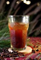 Iced Americano in a large glass with visible refreshing ice cubes, placed on a wooden coaster. Coffee beans are scattered on colorful fabric, and the background shows bokeh lights and green leaves. photo