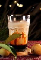 A large glass of palm sugar coffee with an enticing mix of palm sugar syrup and milk. Placed on a small wooden coaster with background with bokeh lights and green foliage. photo