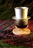 Vietnamese drip coffee in a glass with a metal filter on a wooden coaster, surrounded by coffee beans. The background features bokeh lights and green leaves, providing a calm and natural ambiance. photo