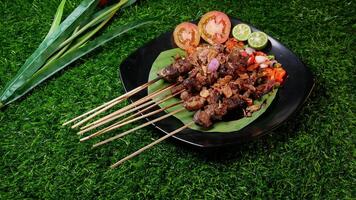 Juicy goat satay with soy sauce and fried shallots, accompanied by fresh tomato sambal, lime slices, and tomato on a banana leaf on a black plate. photo