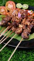 Traditional goat satay with soy sauce, sprinkled with fried shallots, served with fresh tomato sambal, lime slices, and tomato on a black plate with a banana leaf. photo