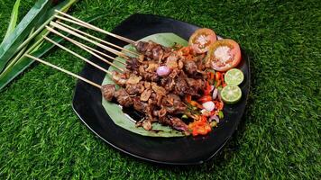 Tender goat satay with soy sauce, sprinkled with fried shallots, served with fresh tomato sambal, lime slices, and tomato on a banana leaf on a black plate. photo
