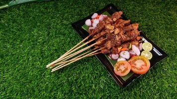 Flavorful chicken satay drenched in peanut sauce, topped with crispy fried shallots, accompanied by fresh tomatoes, shallots, chilies, and lime slices on a banana leaf over a black plate. photo