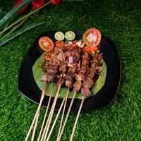 Delicious goat satay with soy sauce, garnished with fried shallots, served with fresh tomato sambal, lime slices, and tomato on a banana leaf. photo
