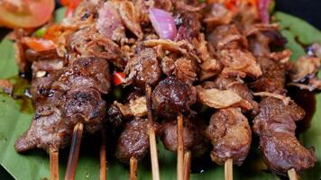 Tender goat satay with soy sauce, sprinkled with fried shallots, served with fresh tomato sambal, lime slices, and tomato on a black plate. photo