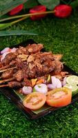Traditional chicken satay with peanut sauce and a sprinkle of fried shallots, served with fresh tomatoes, shallots, chilies, and lime on a banana leaf. photo