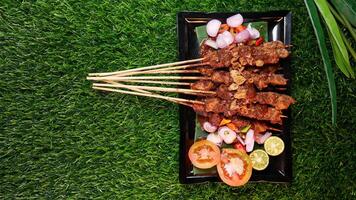 Flavorful chicken satay with peanut sauce, garnished with fried shallots, accompanied by fresh tomatoes, shallots, chilies, and lime slices on a banana leaf over a black plate. photo