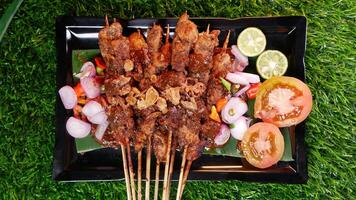 Juicy chicken satay with a generous peanut sauce, topped with fried shallots, served alongside fresh tomato slices, shallots, chilies, and lime on a black plate with a banana leaf. photo