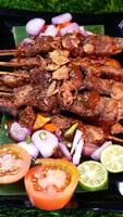 Tasty chicken satay with peanut sauce, garnished with fried shallots, served with fresh tomatoes, shallots, chilies, and lime slices on a banana leaf over a black plate. photo