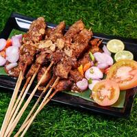 Delicious chicken satay topped with fried shallots and peanut sauce, served with sliced shallots, chilies, fresh tomatoes, and lime on a banana leaf over a black plate. photo