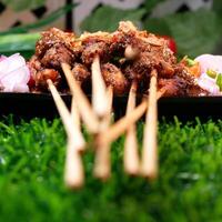 Tasty chicken satay with peanut sauce, garnished with fried shallots, served with fresh tomatoes, shallots, chilies, and lime slices on a banana leaf over a black plate. photo