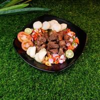 Delicious grilled goat sprinkled with fried shallots and red onions, served with rice cakes, fresh tomato slices, and lime on a banana leaf over a black plate. photo