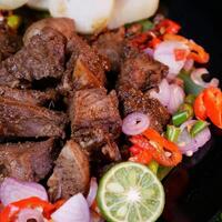 Perfectly seasoned grilled goat sprinkled with fried shallots and red onions, accompanied by rice cakes, lime slices, and fresh tomatoes on a banana leaf. photo