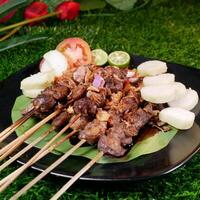 Delicious goat satay sprinkled with fried shallots and red onions, served with rice cakes, fresh tomatoes, and lime on a black plate with a banana leaf. photo