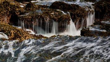 The crashing waves at Karang Bokor Sawarna Beach flowing over the coral rocks become a mini waterfall. The rough texture of the coral rocks and green moss add dimension to the scenery. photo