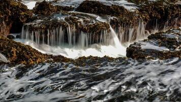 The crashing waves at Karang Bokor Sawarna Beach flowing over the coral rocks become a mini waterfall. The rough texture of the coral rocks and green moss add dimension to the scenery. photo