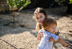 Playful siblings outdoors in a sunny park setting, an older sister lovingly embraces and supports her younger brother, capturing a moment of care, warmth, and family bonding in nature's backdrop. photo