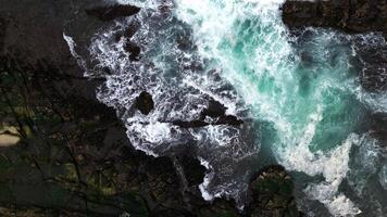The ocean waves crash against the black rocks, creating white foam that contrasts with the dark green seawater. The water flow appears strong, breaking through the rough rock crevices. photo