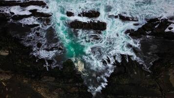 The ocean waves crash against the black rocks, creating white foam that contrasts with the dark green seawater. The water flow appears strong, breaking through the rough rock crevices. photo