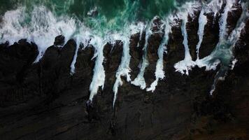 The ocean waves crash against the black rocks, creating white foam that contrasts with the dark green seawater. The water flow appears strong, breaking through the rough rock crevices. photo
