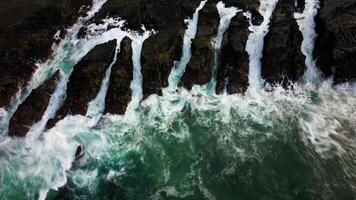 The ocean waves crash against the black rocks, creating white foam that contrasts with the dark green seawater. The water flow appears strong, breaking through the rough rock crevices. photo