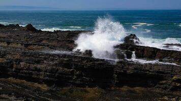 Strong ocean waves crash against solid coral rocks, creating beautiful high water sprays under the bright blue sky and vast ocean at Karang Taraje Beach, Sawarna, Indonesia. photo