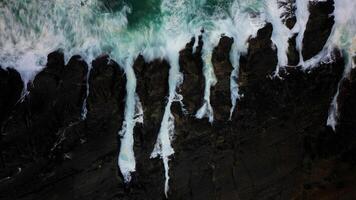 The ocean waves crash against the black rocks, creating white foam that contrasts with the dark green seawater. The water flow appears strong, breaking through the rough rock crevices. photo