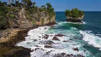 An exotic view of Karang Bokor Beach with high and green rock cliffs, surrounded by clear blue seawater. The waves crashing against the rocks create a beautiful photo