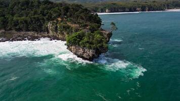 An aerial photo of Karang Bokor Beach displaying spectacular rock formations with green trees on top, surrounded by clear blue seawater and crashing waves.