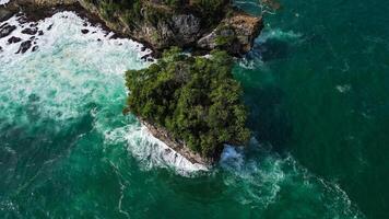 An aerial photo of Karang Bokor Beach displaying spectacular rock formations with green trees on top, surrounded by clear blue seawater and crashing waves.
