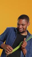 Vertical Happy person having fun playing guitar at a leek in studio, acting funny and mimicking a green onion as a musical instrument. Young guy being silly with organic vegetables. Camera B. video