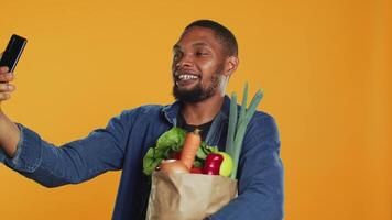 Vegan person talking on videocall conference while shopping for groceries, presenting chemicals free natural fruits and veggies in a paper bag. Young guy recommends local farming. Camera A. video