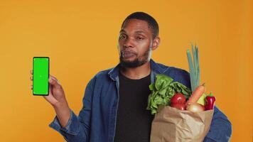 Young guy with a bag of fresh groceries showing greenscreen on his smartphone, presenting an isolated display on phone. Person advertising locally grown produce, zero waste. Camera A. video