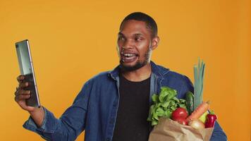 Cheerful guy recommends bio homegrown goods on call, using a tablet to present his groceries from the zero waste eco friendly store. Vegan person talks on teleconference. Camera A. video