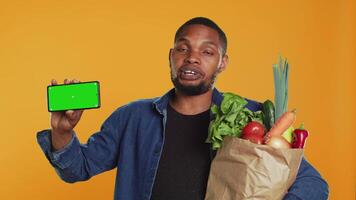 Male model showing a copyspace isolated screen in studio, advertising freshly harvested fruits and veggies from local farmers market. Young guy presenting mockup display on his phone. Camera A. video