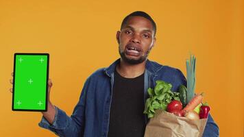 Vegan adult holding a tablet with greenscreen layout in studio, carrying paper bag full of ethically sourced chemicals free produce. Guy advocating for healthy eating with mockup screen. Camera A. video