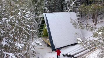 View from the height. Clip. A winter forest in which a man in a red jacket shakes snow off the roof of an unfinished house. video