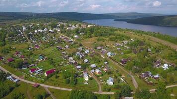 View from the height . Clip . A small summer village with a river, small residential houses and roads, a green forest with large trees and hills is visible from behind. video