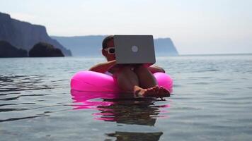 Woman freelancer works on laptop swimming in sea on pink inflatable ring. Happy tourist in sunglasses floating on inflatable donut and working on laptop computer in calm ocean. Remote working anywhere video